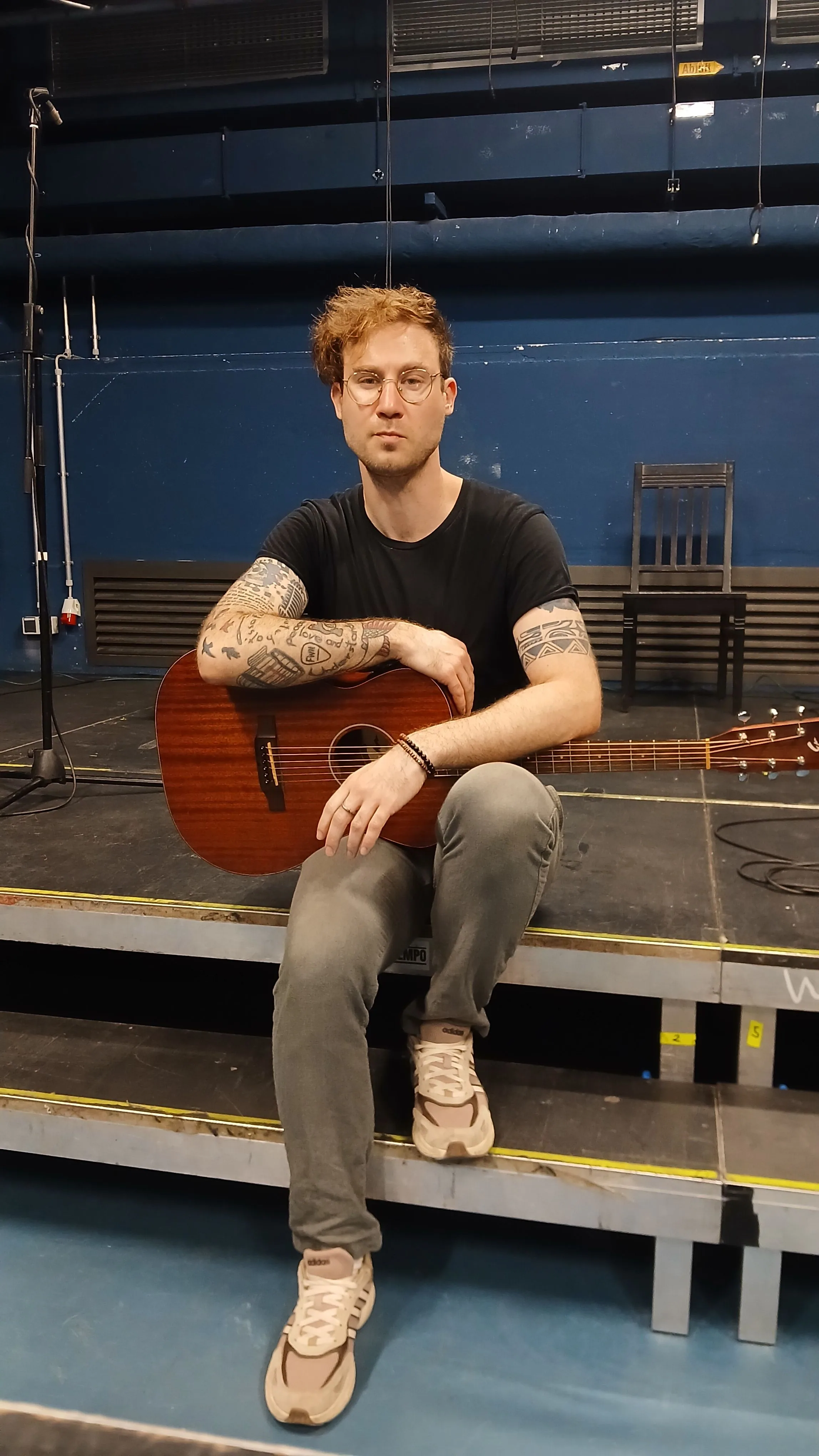 Bricky Waters sitzt mit Akustikgitarre auf einer Bühnenkante vor blauer Wand